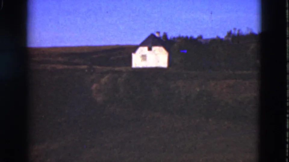1975 Denmark home-movie view of a distant white house on a rural hillside, with open fields and a dark tree line behind it.