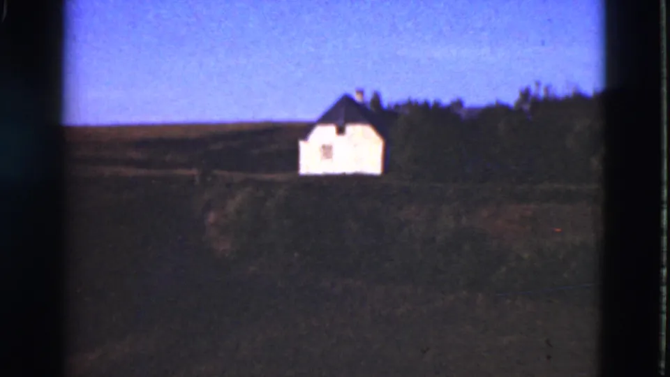 1975 Denmark rural view showing a white house with a dark roof across open fields, framed by trees and a clear blue sky.