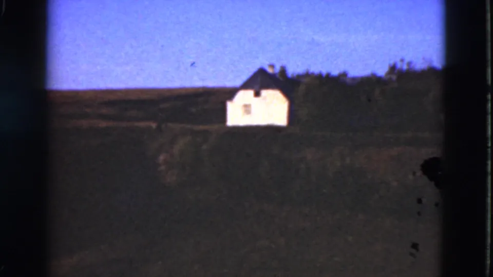 1975 Denmark home-movie frame of a white hillside house, open foreground fields, and trees, with visible film grain and edge shadowing.