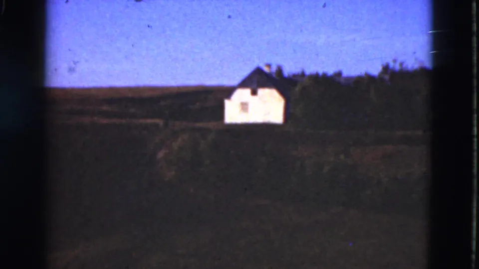 1975 Denmark home-movie frame with a distant white house, dark roof, trees, and open rural ground.