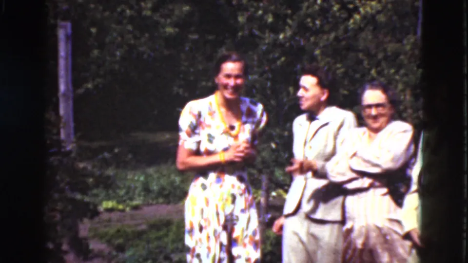 1975 Denmark 8mm frame of two women and a man smiling together in a garden, with one woman in a floral dress.