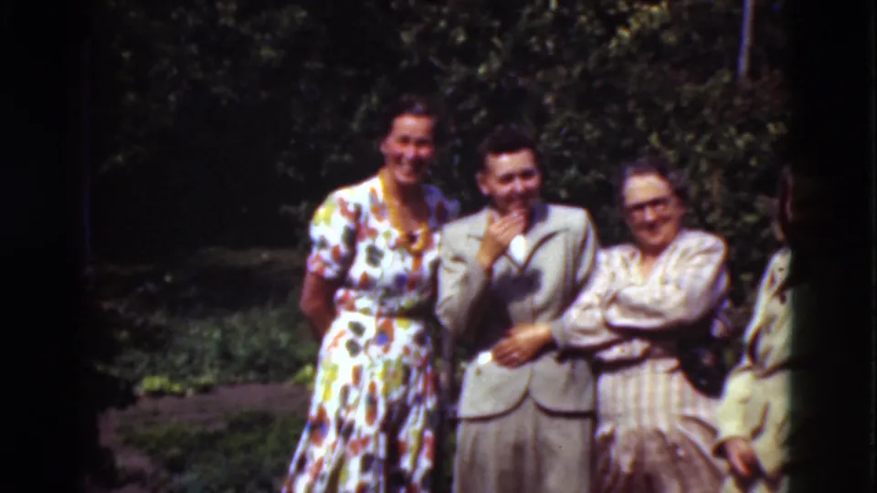 1975 Denmark home-movie frame showing adults smiling in a garden, including a woman in a floral dress and a man in a light suit.