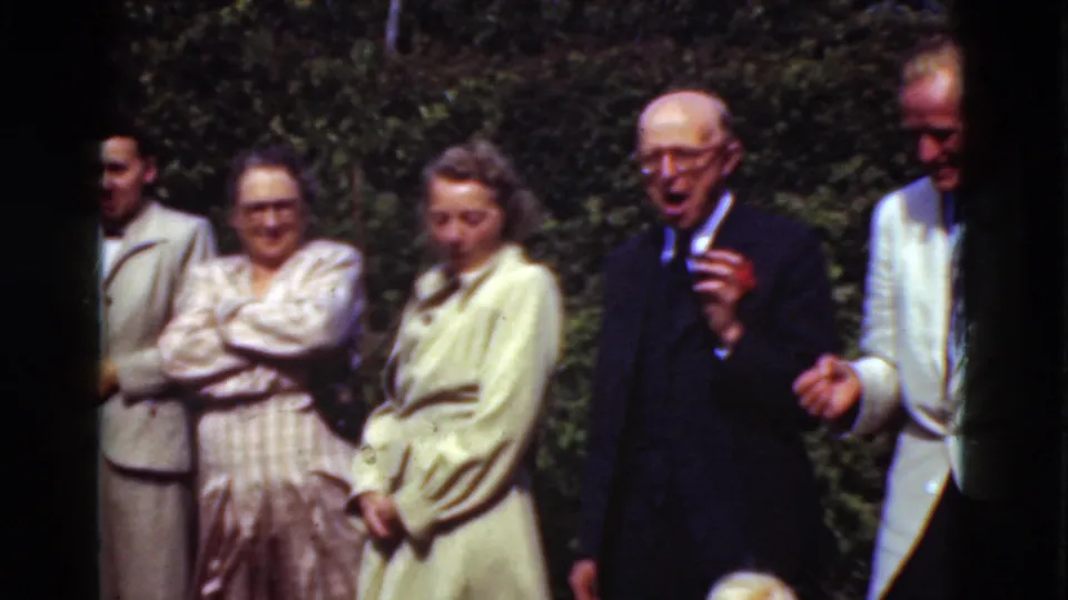 A Denmark 1975 home-movie frame shows adults standing in a garden as an older man in a dark suit appears to talk or laugh.