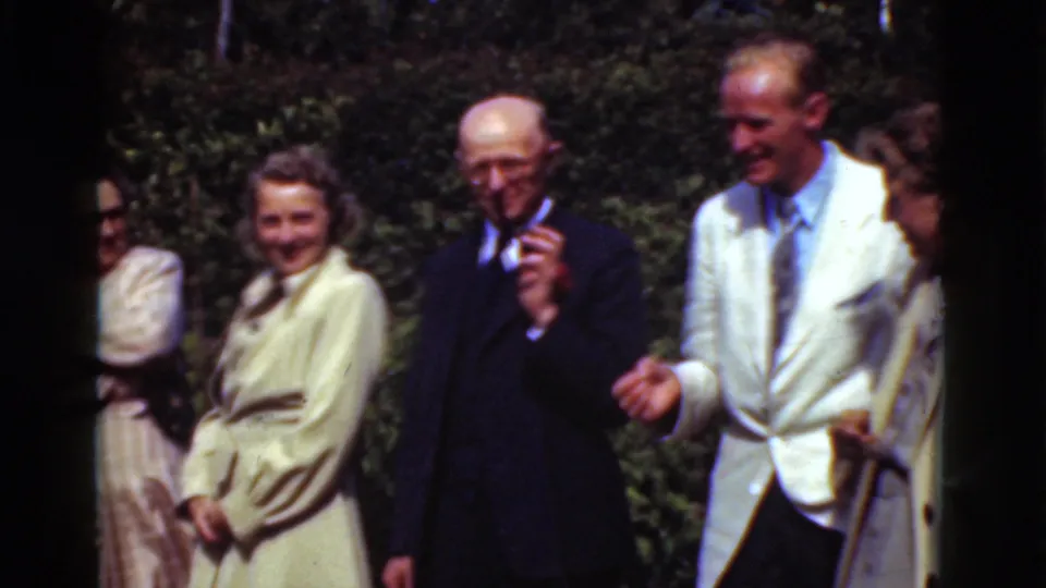 In Denmark 1975 8mm footage, adults in dress clothing stand outdoors, including an older man in a dark suit and a man in a pale jacket.