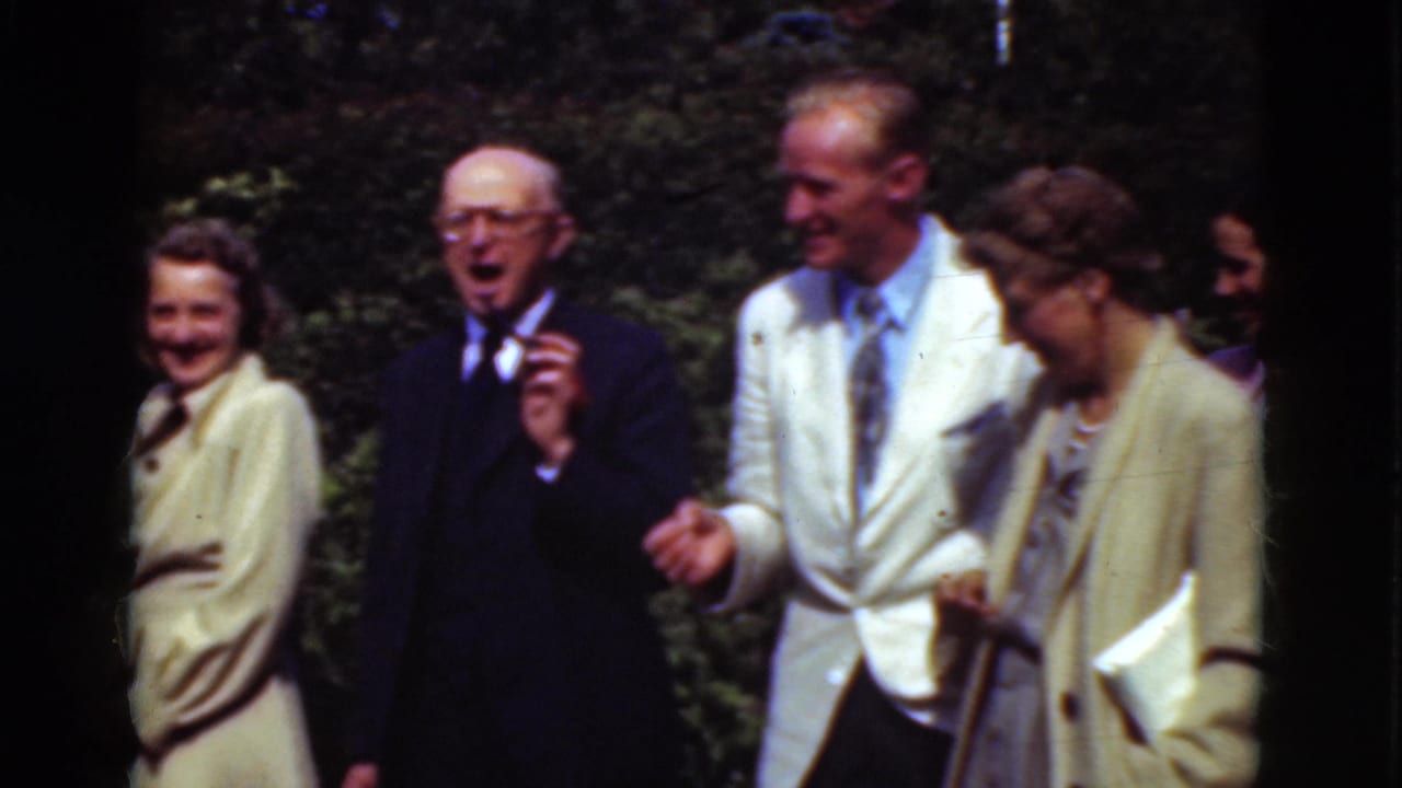 Adults in formal clothing smile and talk outdoors in bright sunlight.