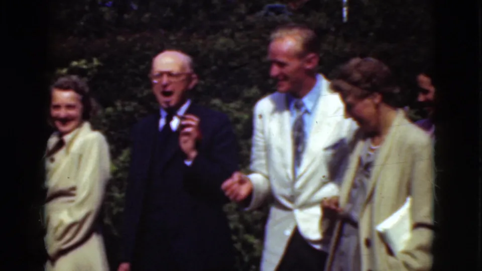 In Denmark archival home-movie footage from 1975, adults in suits and coats stand outdoors smiling near greenery.