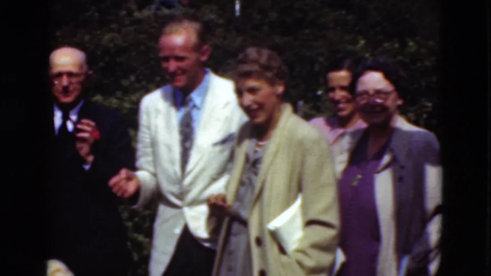 A 1975 Denmark home-movie frame shows a smiling group walking through a sunny outdoor setting in formal clothes.