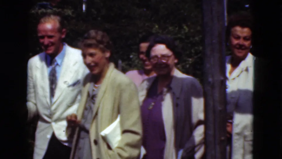 Adults in 1970s formal clothing, including a man in a white jacket and a woman with glasses, move past the handheld camera.