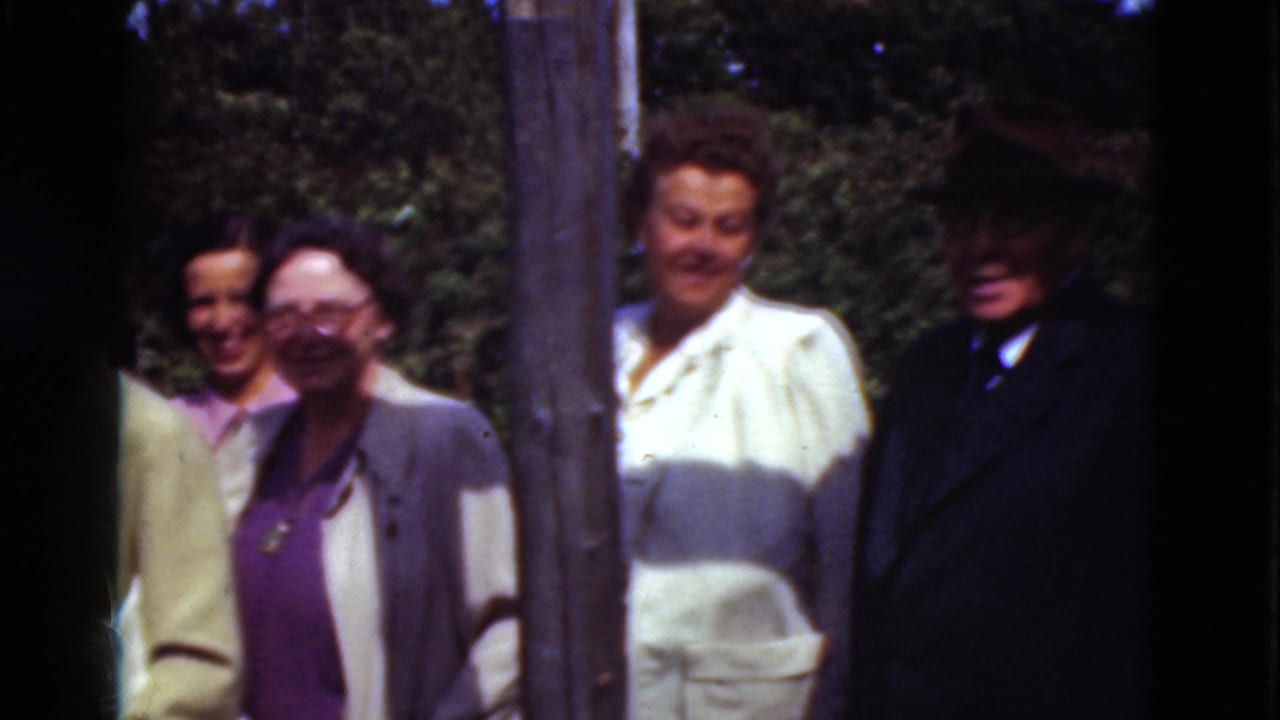 Adults standing outside near a wooden post, hedges and a red-roofed building.