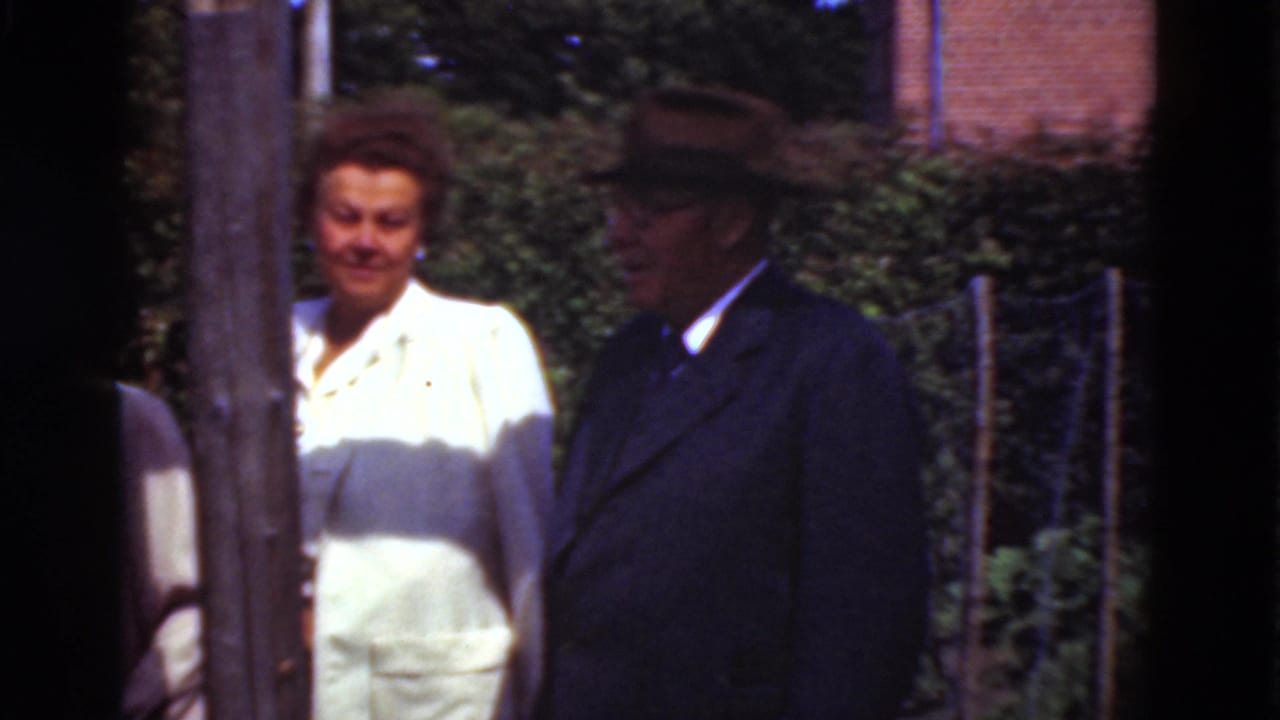 Woman in a light dress and man in dark suit and hat standing beside hedges.