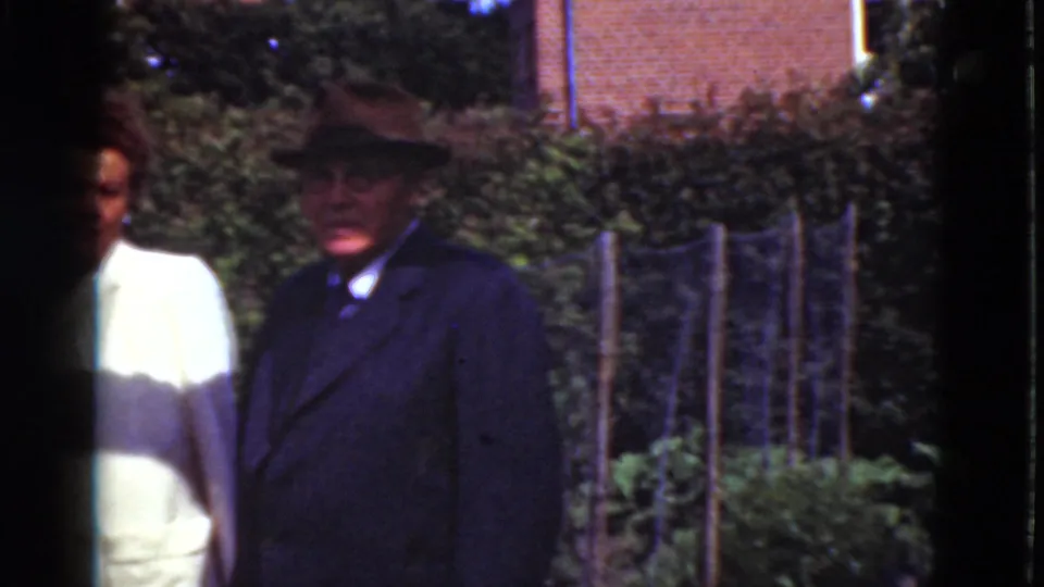 1975 Denmark home-movie still of a suited man in a hat and a woman in a light dress standing by hedges, fencing and a brick building edge.