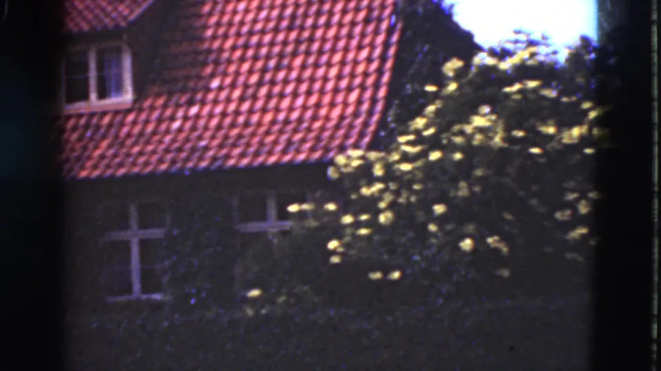 1975 Denmark 8mm frame with a red tiled roof, small dormer, white-framed windows and dense foliage in front of the facade.