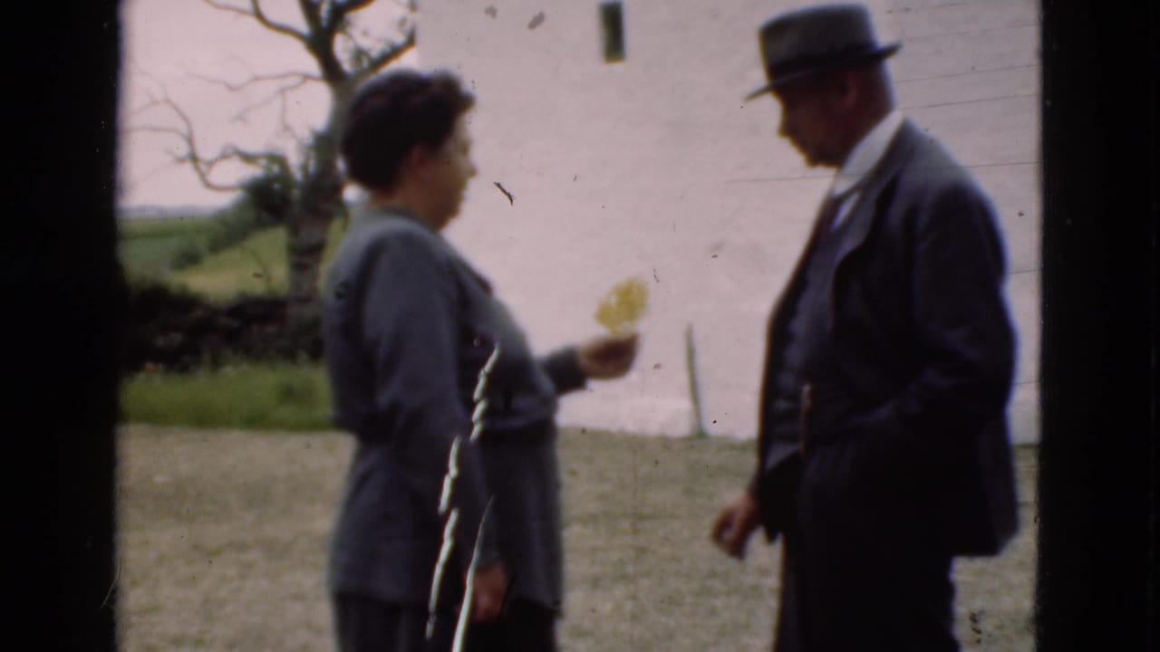 In 1975 Denmark home-movie footage, a woman with a small yellow flower stands near a suited man in a hat beside a white wall.