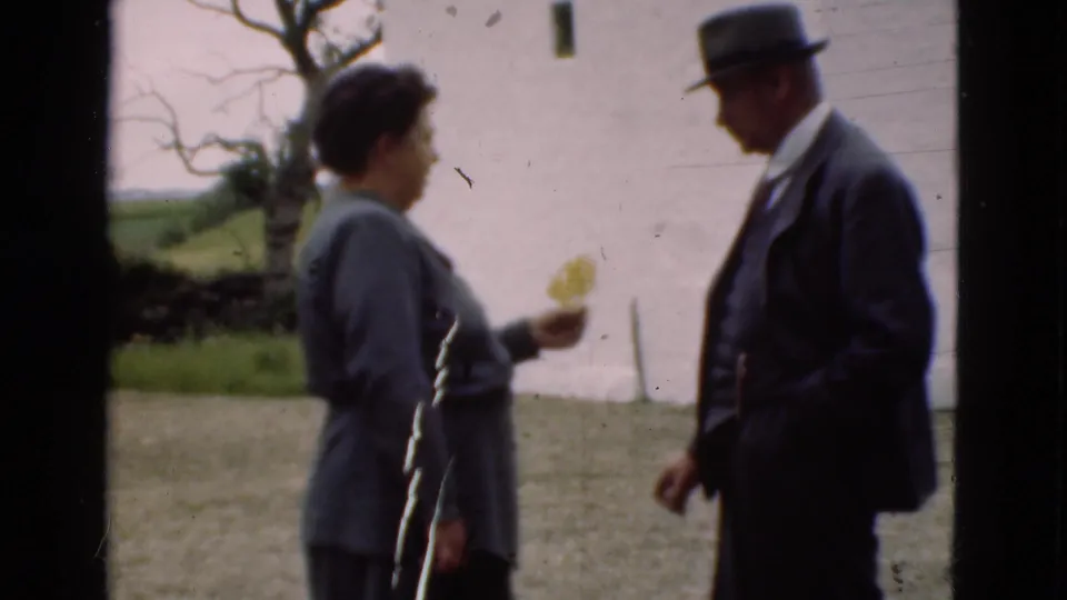 In 1975 Denmark home-movie footage, a woman with a small yellow flower stands near a suited man in a hat beside a white wall.