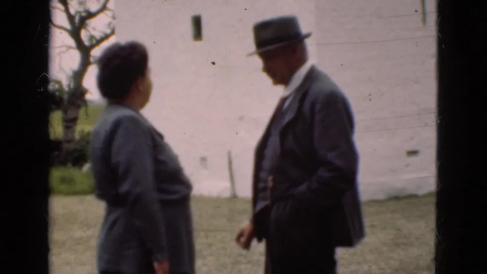 The two older adults face each other in a soft 8mm frame, with a white wall, grass, and rural background visible.
