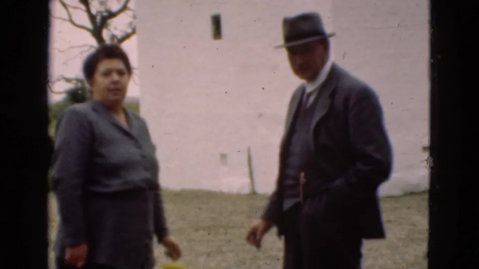 In the final frame, the woman and suited man turn toward the camera in the Denmark backyard scene, with the white wall behind them.