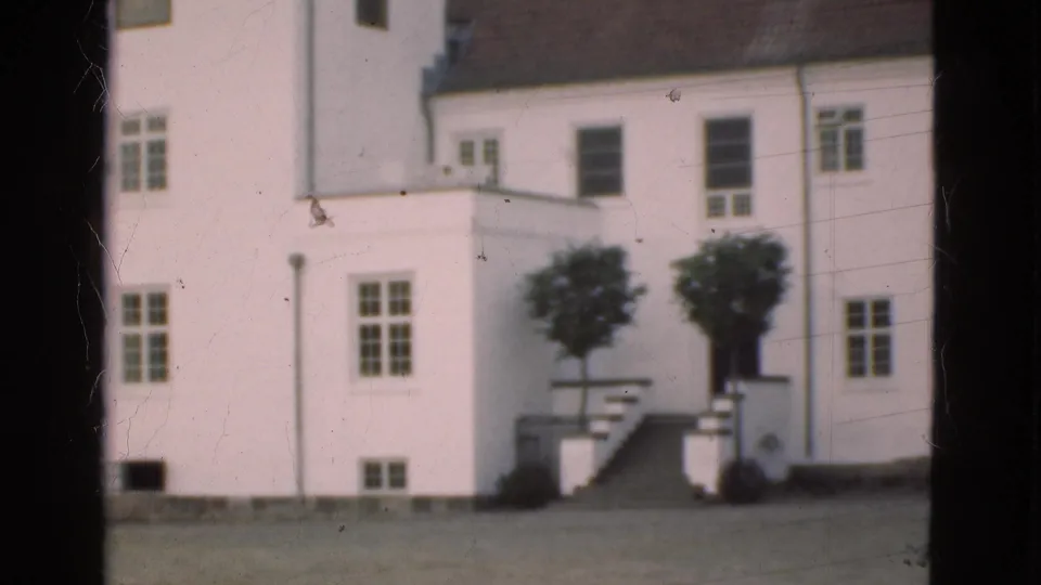 1975 Denmark 8mm frame showing the building corner, windows, exterior steps, trimmed trees, lawn, and visible film scratches.
