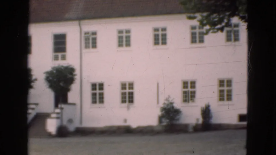 1975 Denmark 8mm frame showing the pan continuing along a white multi-storey facade with stairs at left, windows, and small plantings.