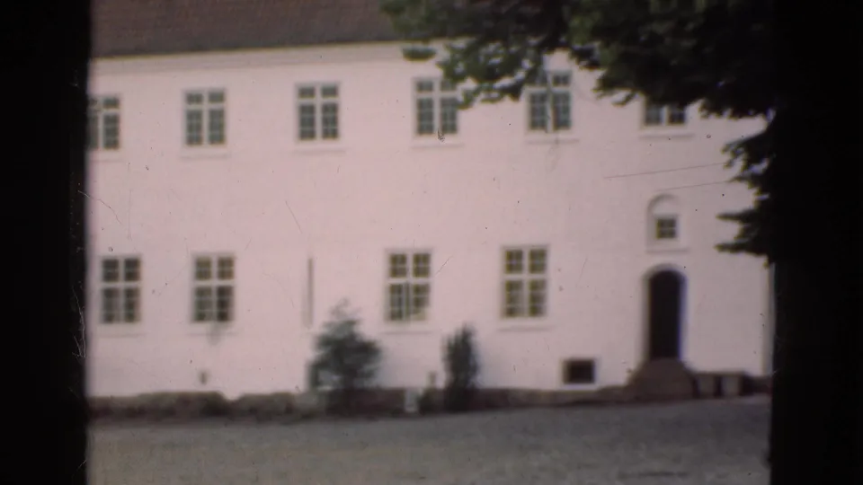 1975 Denmark home-movie frame showing the right side of the white building, including rows of windows, a doorway, shrubs, and overhanging branches.