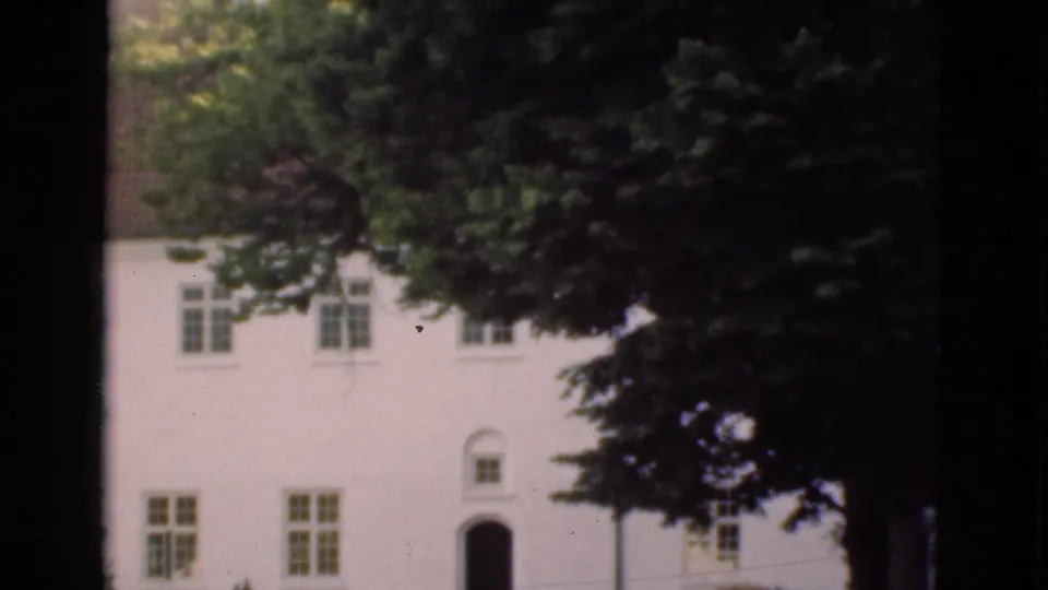 1975 Denmark 8mm frame showing the house facade after the pan, with an arched entrance, upper windows, and dense trees along the right edge.
