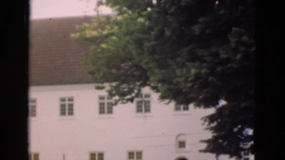 1975 Denmark 8mm frame of the building during the pan, emphasizing the tiled roof, upper windows, white wall, and leafy trees in front.
