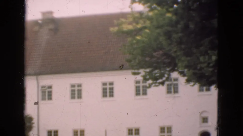 1975 Denmark 8mm frame near the end of the pan, with tiled roof, rows of windows, tree cover, and a dark arched doorway near the right edge.