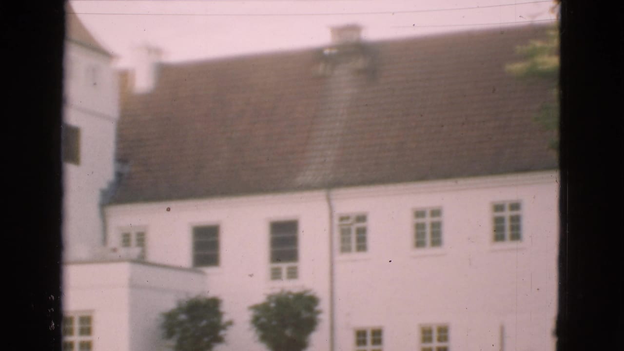1975 Denmark 8mm frame with a white building, brown tiled roof, small windows, trimmed trees, and a dark vertical window frame crossing the view.