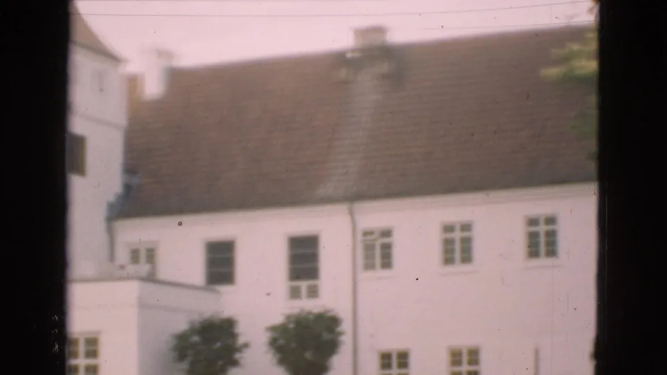 1975 Denmark 8mm frame with a white building, brown tiled roof, small windows, trimmed trees, and a dark vertical window frame crossing the view.