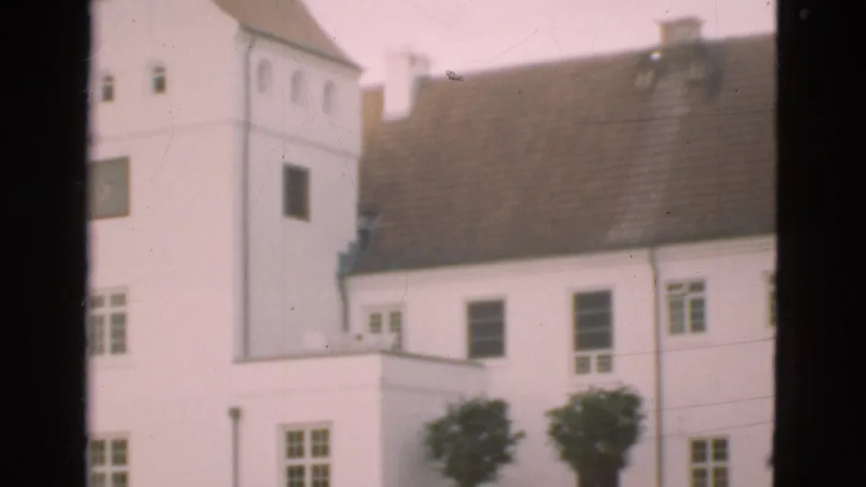 1975 Denmark 8mm frame showing a tower-like white section, brown roof, small windows, foreground window bars, and trees near the base.