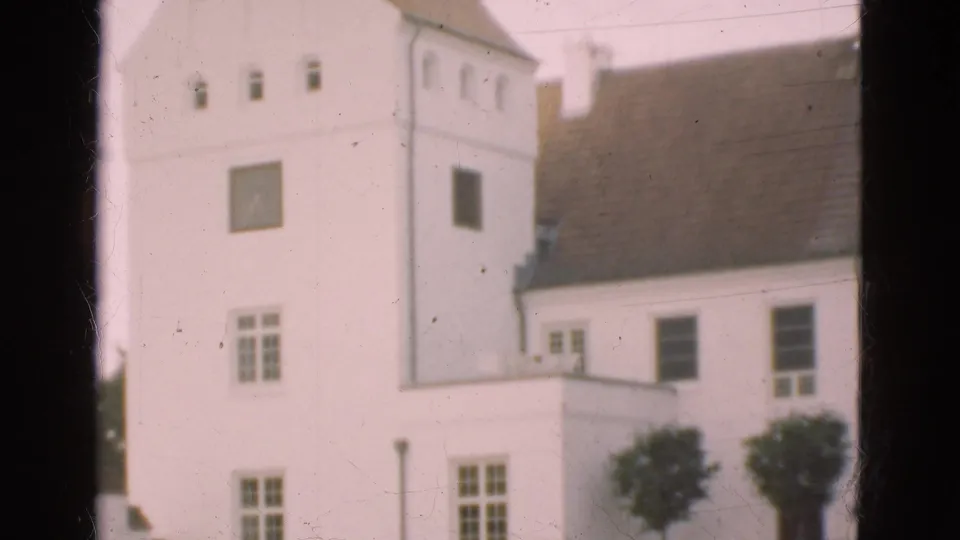 1975 Denmark 8mm frame with a white tower-like building section, roof tiles, small windows, trees, and dark frame edges from the filming position.
