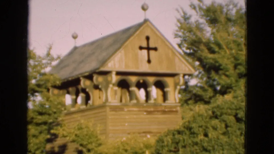 1975 Denmark 8mm frame showing the church tower roof, black cross, decorative finials, arched openings, and green trees.