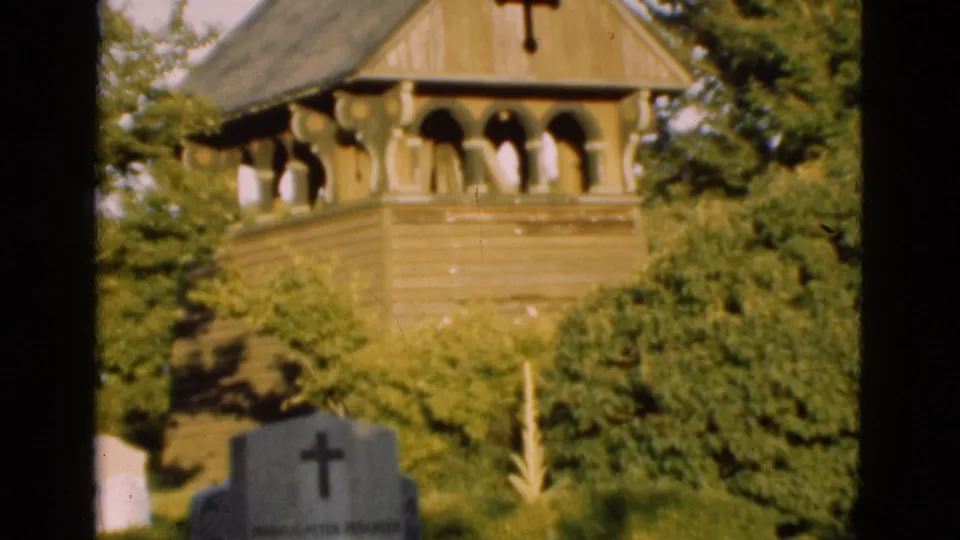 1975 Denmark 8mm frame showing the church tower above dense shrubs and a foreground gravestone marked with a cross.