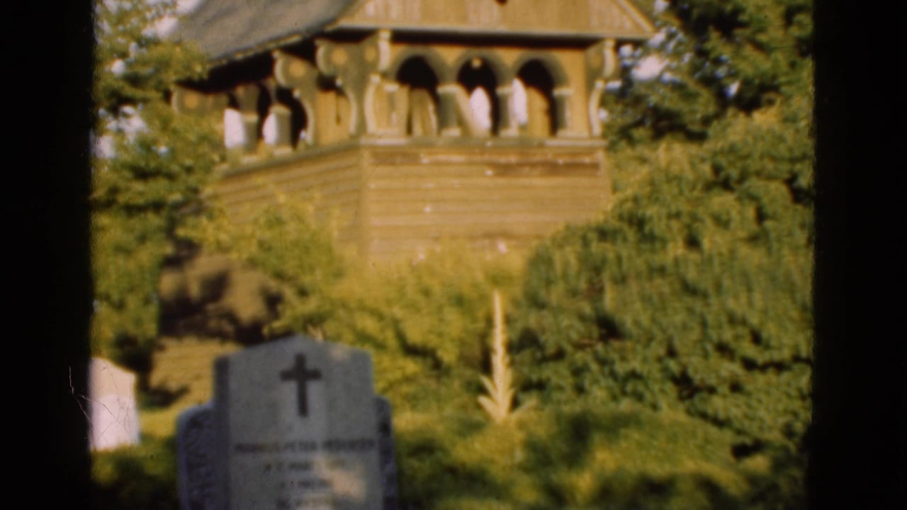 Gravestone with a cross in front of trees and a wooden church tower.