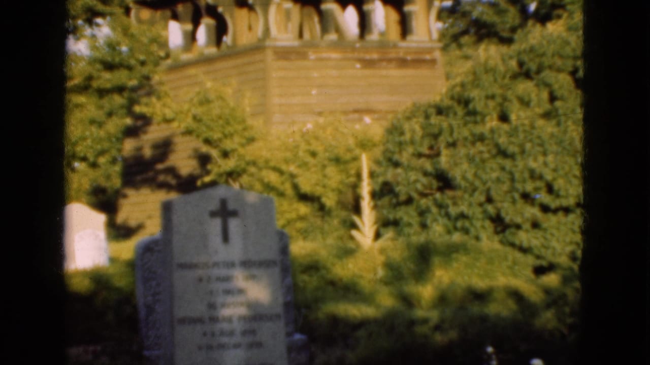 1975 Denmark cemetery view with a cross-topped grave marker, leafy trees, and a wooden church tower seen through dark frame borders.