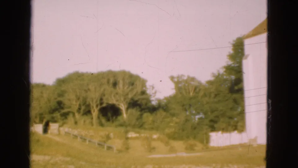 1975 Denmark home-movie frame showing a pale church wall beside grass, trees, and a low fence or path line in the churchyard.
