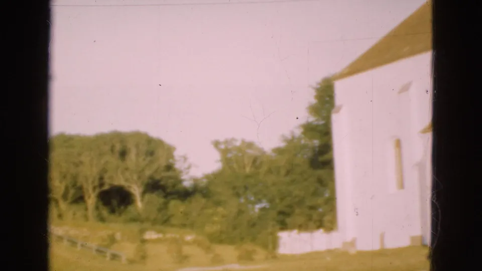 1975 Denmark churchyard frame with the pale church wall at right, grass, trees, and a low fence line under warm faded color.