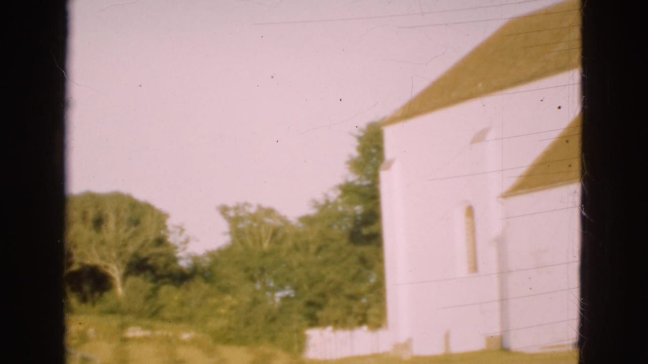 1975 Denmark 8mm frame showing a white church wall, yellow-tiled roof, surrounding trees, grass, and pale sky within dark film borders.