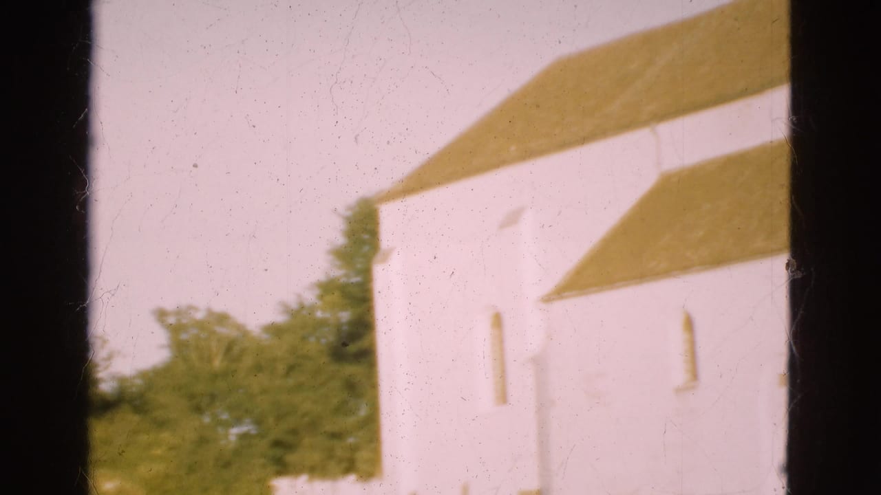 1975 Denmark 8mm frame with the church shifted right, showing the yellow roofline, white wall, arched window, trees, and dark gate edges.