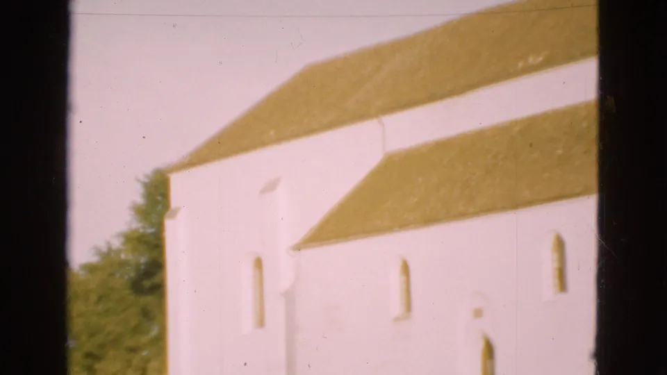 1975 Denmark 8mm frame showing a closer, softer view of the white church exterior with long yellow roof sections, small windows, trees, and gate borders.