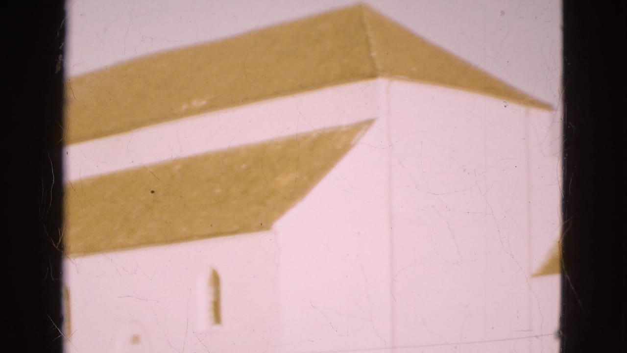 1975 Denmark home-movie frame with a white wall, brown roof edge, arched window and visible film gate border.