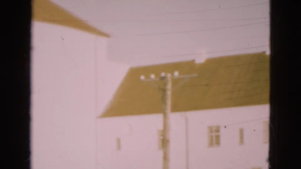 1975 Denmark 8mm frame with a white building side, brown roof, small windows, overhead wires and a utility pole.