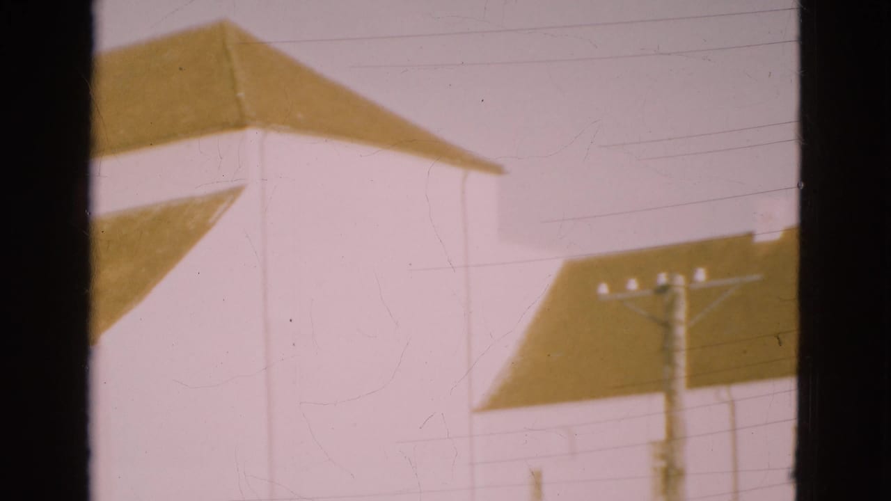 White buildings with brown pitched roofs seen past a dark vertical window frame.