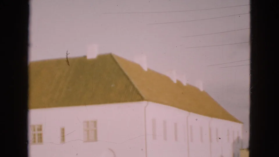 1975 Denmark home-movie frame of a white farmhouse with a green roof, seen in soft focus with dark film-strip borders.