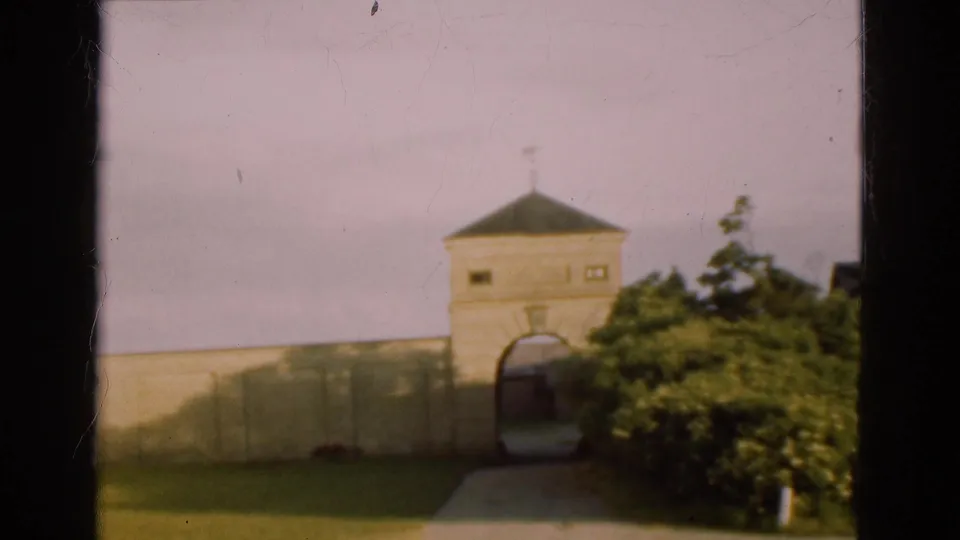 1975 Denmark frame showing a pale stone gatehouse with an arched opening, wall, shrubs, lawn, and walkway.