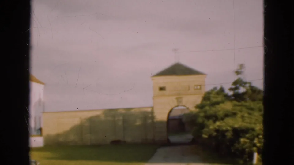 Soft 8mm frame of a Danish village gatehouse with arched entry, pale wall, shrubs, lawn, walkway, and a partial vehicle at left.