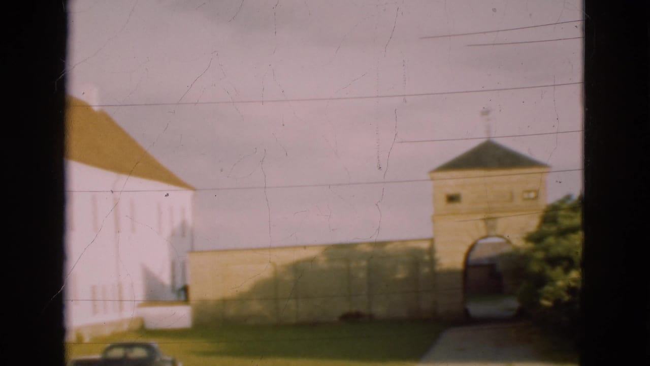 White building with brown roof, stone wall, arched gate, grass, and dark parked car.