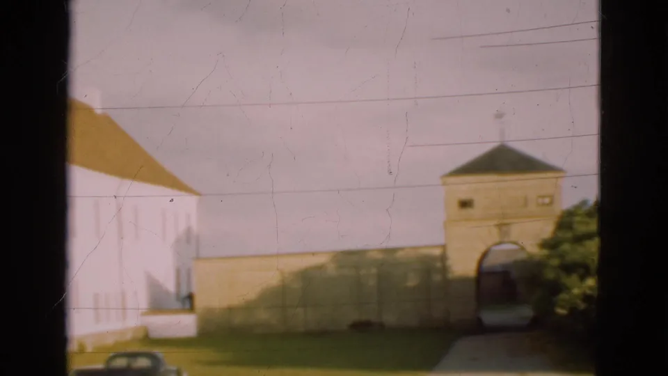 1975 Denmark 8mm frame showing a white building with brown roof, a stone gate and wall, grass, and a dark car at the lower edge.