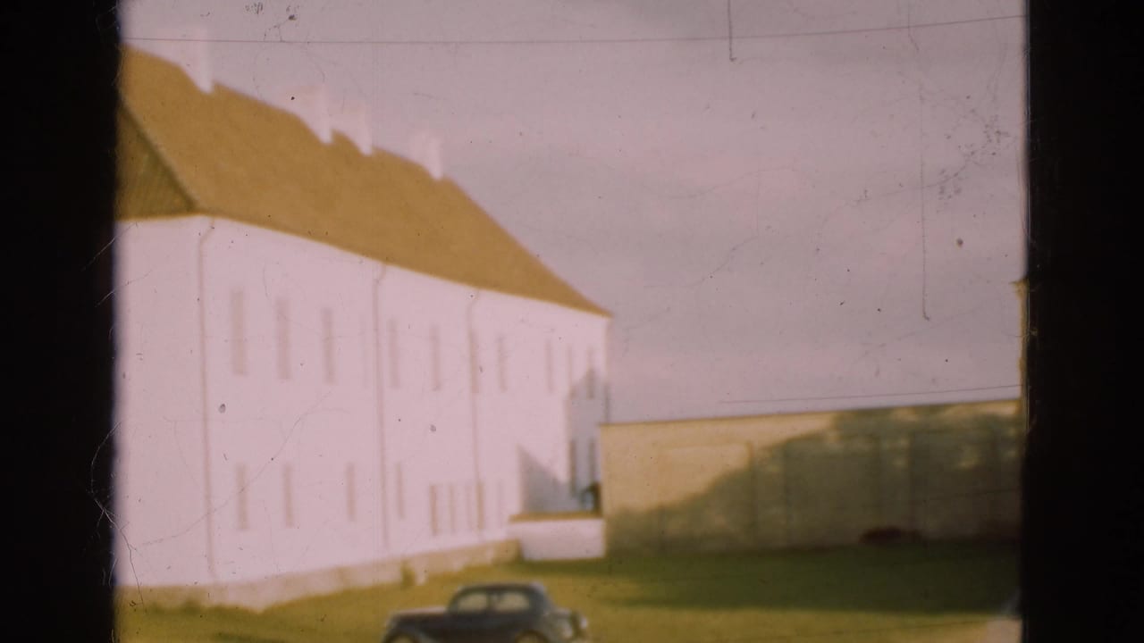 1975 Denmark 8mm frame looking along the white building facade, with a brown roof, grass foreground, wall, and dark parked car.