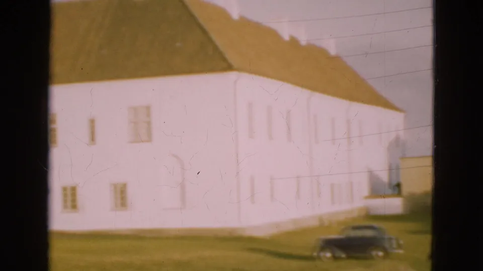 1975 Denmark 8mm frame with a closer view of the white building, brown roof, small windows, grass, and a dark car near the bottom.