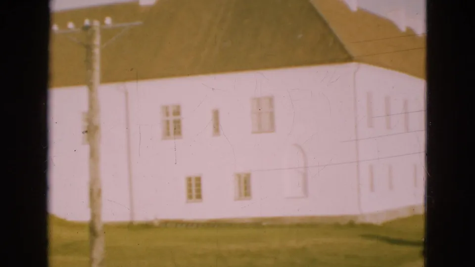 1975 Denmark 8mm frame showing the white building facade with a brown roof, utility pole in front, grass, small windows, and sky.